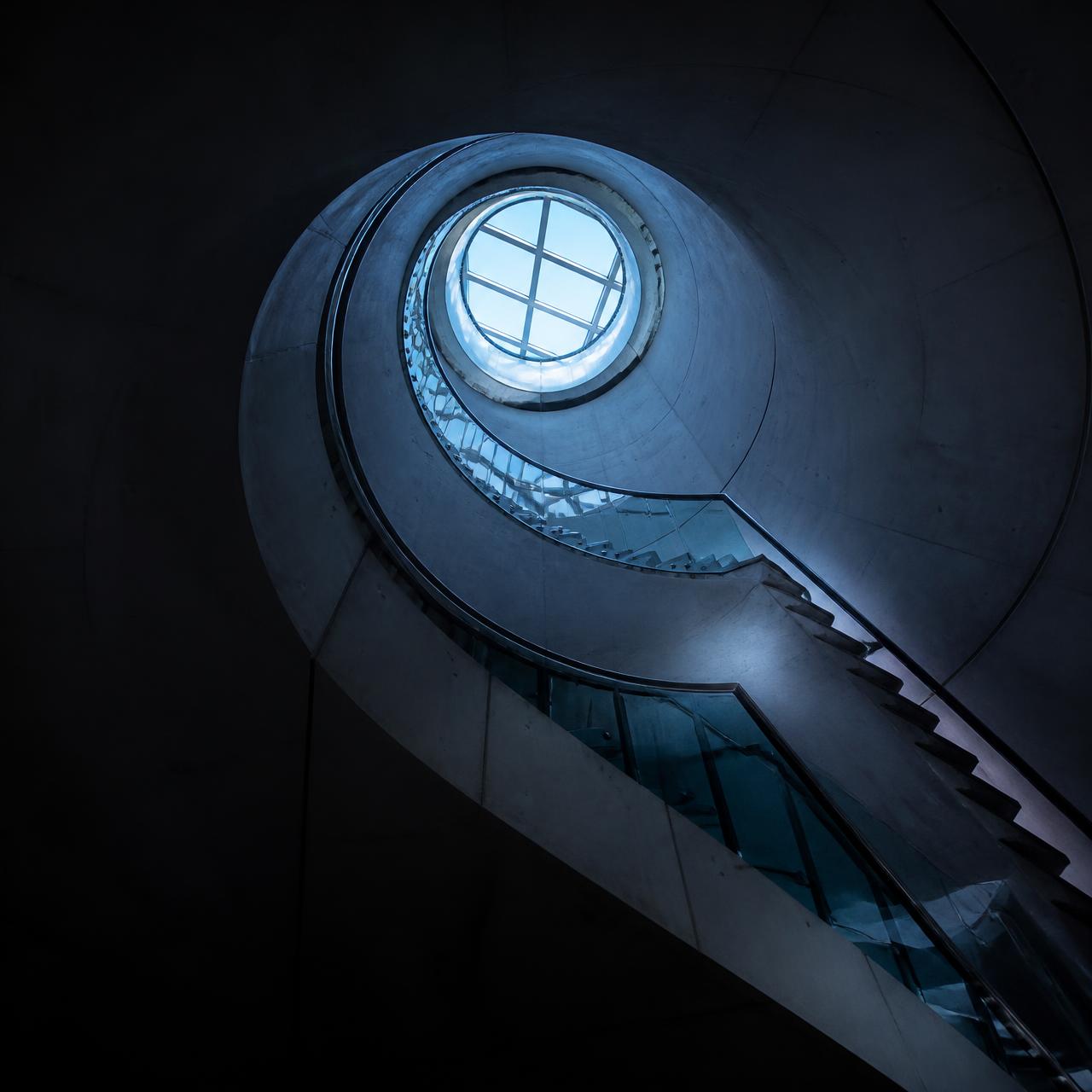 Spiral concrete stairwell rising towards a circular skylight